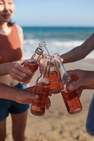 Téléchargez les photos : Image en gros plan d'amis qui trinquent avec des bouteilles en verre lorsqu'ils passent du temps sur la plage - en image libre de droit