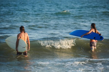 Téléchargez les photos : Surfeurs marchant dans l'océan pour attraper des vagues dans les rayons au coucher du soleil - en image libre de droit