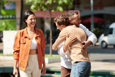 Téléchargez les photos : Femme souriante regardant son petit ami heureux embrasser son meilleur ami qu'il a rencontré dans le parc - en image libre de droit