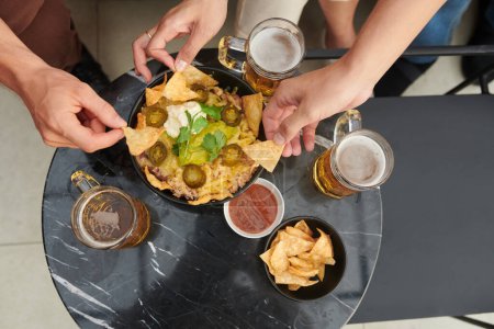 Téléchargez les photos : Mains de personnes buvant des bières avec bol de chips de nacho au riz, tranches de jalapeno, guacamole et crème sure, vue d'en haut - en image libre de droit