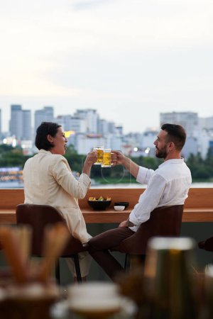 Téléchargez les photos : Amis griller avec des tasses à bière lors du repos dans le bar sur le toit avec une belle vue - en image libre de droit