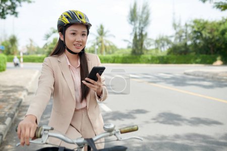 Téléchargez les photos : Portrait d'une femme d'affaires vietnamienne positive enregistrant un message vocal lorsqu'elle se rend au travail à vélo - en image libre de droit