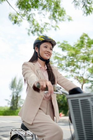Foto de Sonriente joven empresaria asiática en traje beige montando bicicleta a la oficina - Imagen libre de derechos