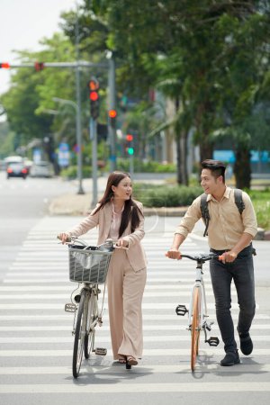 Photo for Positive young Vietnamese businesssman and businessswoman with bicycles crossing road - Royalty Free Image
