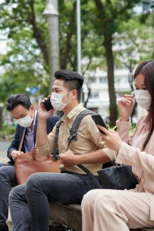 Photo for Frowning young Asian man in protective mask talking on phone when sitting on bench outdoors - Royalty Free Image