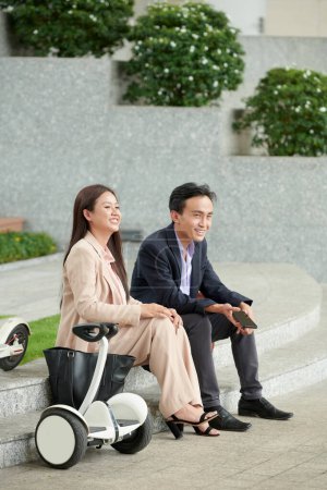 Photo for Happy Vietnamese entrepreneurs sitting on steps outdoors and listening to voice message on smartphone from coworker - Royalty Free Image