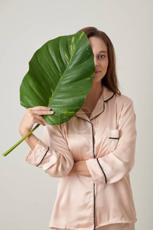 Photo for Portrait of smiling young woman covering half of her face with big lush leaf, and looking at camera - Royalty Free Image