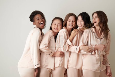 Foto de Mujeres jóvenes positivas en pijamas de satén riéndose y abrazándose - Imagen libre de derechos
