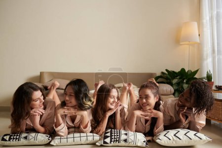 Photo for Positive young women in satin pajamas lying on floor and smiling at each other when enjoying sleepover - Royalty Free Image