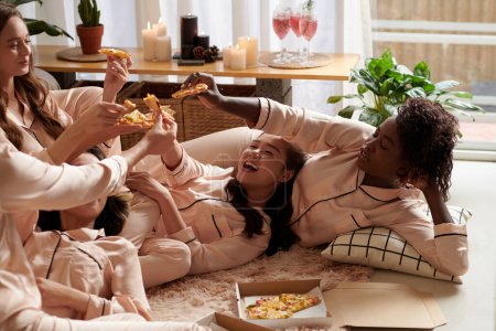 Gruppe junger Frauen prostet sich im Schlafanzug auf dem Boden liegend mit Pizzaschnitten zu