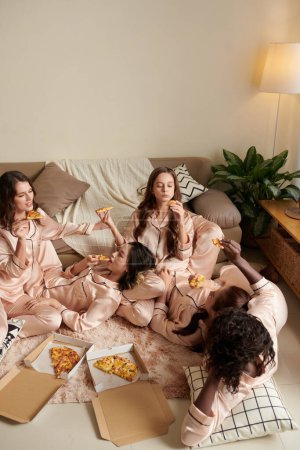 Foto de Grupo de mujeres jóvenes en pijama comiendo pizza y disfrutando de la fiesta de pijamas en casa de amigos - Imagen libre de derechos