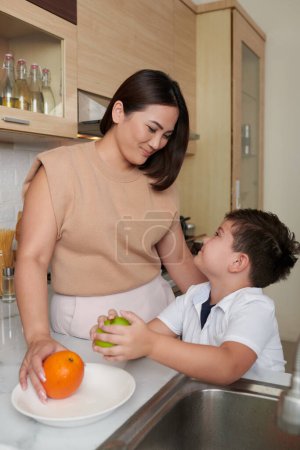 Photo for Happy mother and son putting washed fruits on plate to serve for breakfast - Royalty Free Image
