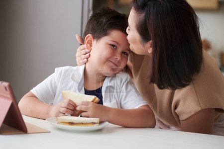 Photo for Mother kissing smiling son on forehead when he is eating sandwiches at kitchen counter - Royalty Free Image