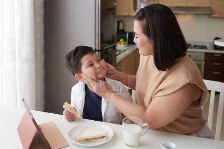 Foto de Madre aplastando las mejillas gordita de su hijo cuando están desayunando en la cocina - Imagen libre de derechos