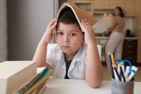 Foto de Triste cabeza de niño cubierta con libro abierto cuando se sienta en la mesa de la cocina - Imagen libre de derechos