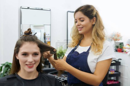 Téléchargez les photos : Coiffeur utilisant une brosse ronde pour coiffer les cheveux d'une cliente souriante - en image libre de droit