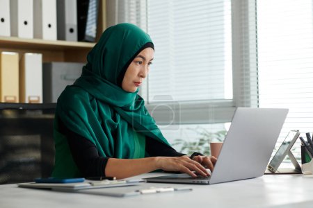 Foto de Mujer musulmana seria en hijab trabajando en la computadora portátil en la oficina - Imagen libre de derechos