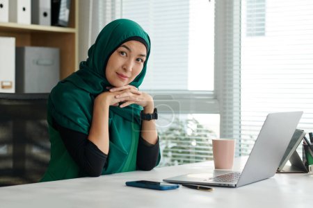 Photo for Portrait of smiling muslim female entrepreneur sitting at office desk and looking at camera - Royalty Free Image