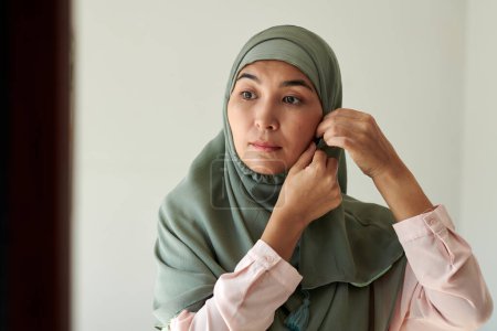 Foto de Retrato de mujer musulmana madura poniéndose hiyab de seda teal - Imagen libre de derechos