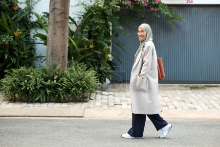 Téléchargez les photos : Femme musulmane souriante en manteau de laine et hijab marchant dans la rue - en image libre de droit