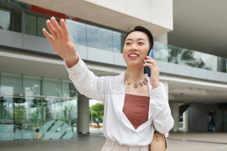 Téléchargez les photos : Joyeuse femme d'affaires asiatique parlant au téléphone et attrapant taxi voiture - en image libre de droit