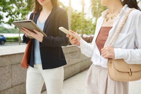Foto de Imagen recortada de mujeres de negocios caminando en la calle y comprobando mensajes en la tableta y el teléfono inteligente - Imagen libre de derechos