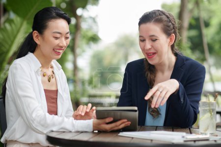Foto de Mujeres de negocios alegres sentados en la mesa en la cafetería al aire libre y discutir el contrato en la computadora tableta - Imagen libre de derechos