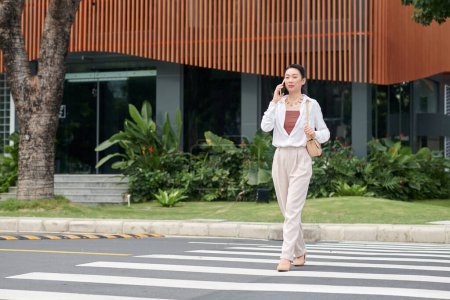 Foto de Joven mujer de negocios vietnamita hablando por teléfono y cruzando la calle en la ciudad - Imagen libre de derechos