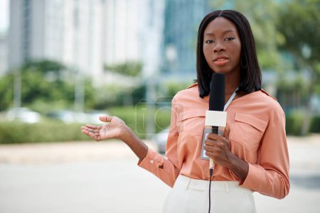 Téléchargez les photos : Journaliste noir confiant parlant dans le microphone à l'extérieur lors du tournage du nouveau rapport - en image libre de droit