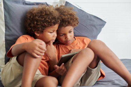 Foto de Hermanos sentados en la cama de los padres y viendo dibujos animados en la tableta - Imagen libre de derechos