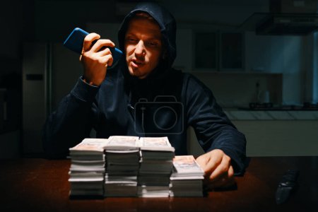 Téléchargez les photos : Voleur assis à table dans la pièce sombre, regardant pile d'argent et enregistrement message vocal - en image libre de droit