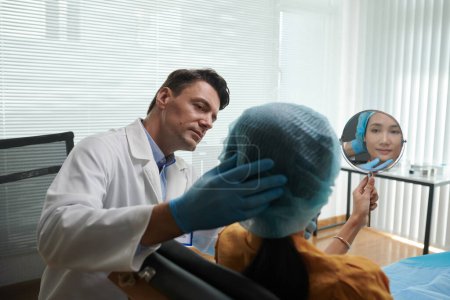 Téléchargez les photos : Chirurgien plastique laissant le patient regarder dans le miroir après la thérapie injections - en image libre de droit