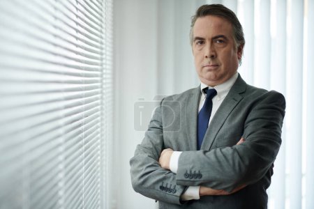 Téléchargez les photos : Portrait d'un homme d'affaires sérieux et confiant debout à la fenêtre du bureau les bras croisés - en image libre de droit
