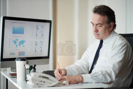 Photo for Concentrated mature entrepreneur analyzing chart on computer screen and taking notes - Royalty Free Image