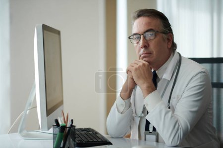 Foto de Retrato del médico general serio que trabaja en la computadora en su escritorio - Imagen libre de derechos
