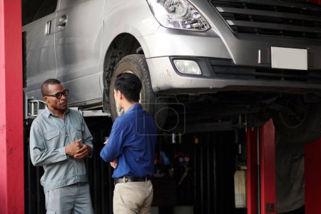 Téléchargez les photos : Propriétaire de voiture et mécanicien debout à la voiture suspendue et discuter des détails de la réparation automobile - en image libre de droit
