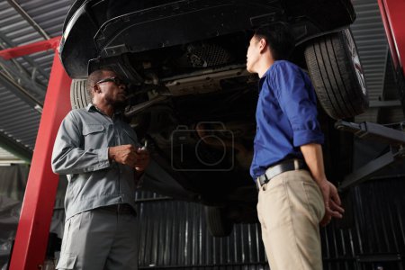 Téléchargez les photos : Propriétaire de voiture et mécanicien debout sous la voiture suspendue et discuter de ce qui doit être fixé - en image libre de droit