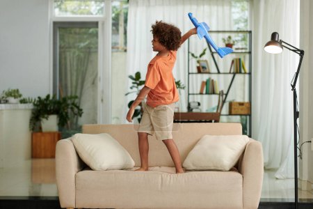 Foto de Niño de pie en el sofá y jugando con el avión de juguete imaginándose a sí mismo como un piloto - Imagen libre de derechos