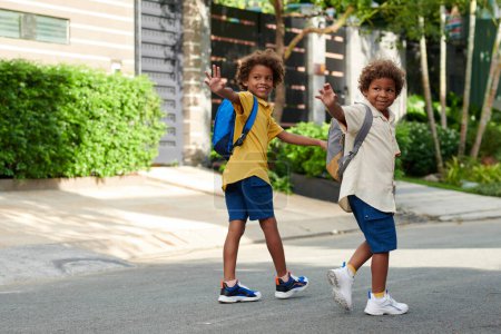 Téléchargez les photos : Des enfants joyeux saluent leurs parents en quittant la maison et en allant à l'école à pied - en image libre de droit