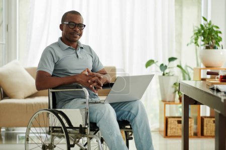 Téléchargez les photos : Portrait d'un homme noir souriant avec un ordinateur portable assis en fauteuil roulant dans son appartement - en image libre de droit