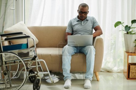 Téléchargez les photos : Homme handicapé assis sur un canapé à côté d'un fauteuil roulant et travaillant sur un ordinateur portable - en image libre de droit