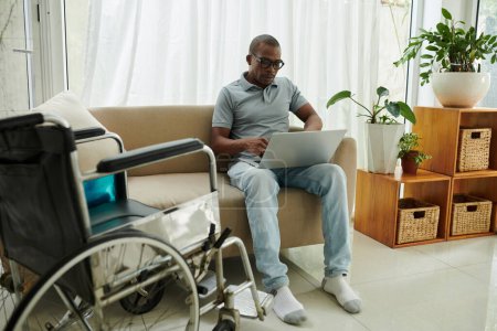 Foto de Hombre recuperándose de lesiones graves sentado en el sofá junto a la silla de ruedas y trabajando en el ordenador portátil - Imagen libre de derechos
