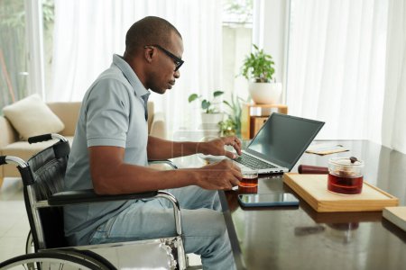 Photo for Black man with disability sitting at desk, drinking tea and checking e-mails on laptop - Royalty Free Image