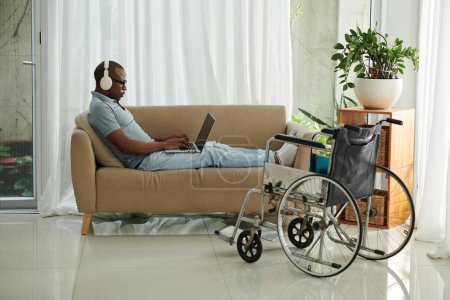 Téléchargez les photos : Homme handicapé assis sur le canapé à la maison et travaillant ou jouant sur un ordinateur portable - en image libre de droit