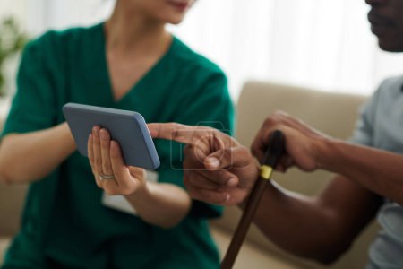 Foto de Enfermera médica visitando al paciente en casa y mostrándole aplicación de telemedicina en smartphone - Imagen libre de derechos