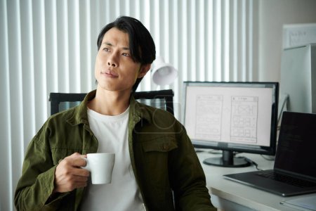 Foto de Retrato de desarrollador de software inspirado pensativo con taza de café sentado en su escritorio - Imagen libre de derechos