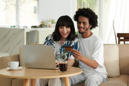Téléchargez les photos : Heureux jeune couple interculturel choisissant des biens en ligne devant un ordinateur portable tandis que l'homme afro-américain avec carte de crédit va payer pour l'achat - en image libre de droit