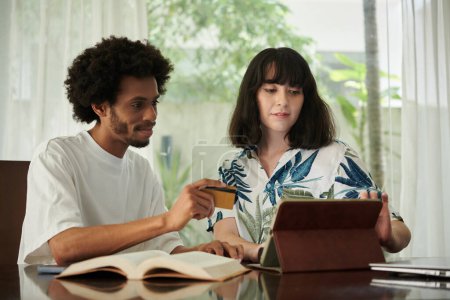 Téléchargez les photos : Jeune homme tenant une carte de crédit et le montrant à sa femme entrant son numéro sur le site alors que les deux assis devant l'écran de la tablette - en image libre de droit