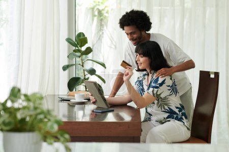 Foto de Feliz joven y su novia con tarjeta de crédito elegir productos en línea y ordenar algunos de ellos mientras se mira la pantalla de la tableta - Imagen libre de derechos