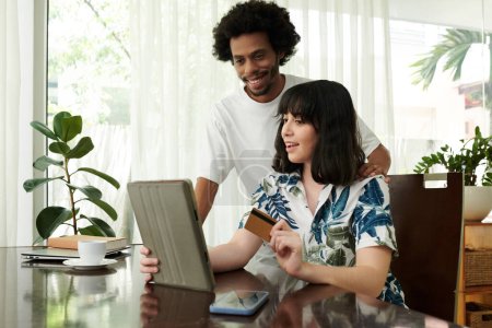 Foto de Mujer joven con tarjeta de crédito y su marido mirando a través de productos en línea en la pantalla de la tableta, elegir algunos y pagar por ellos - Imagen libre de derechos